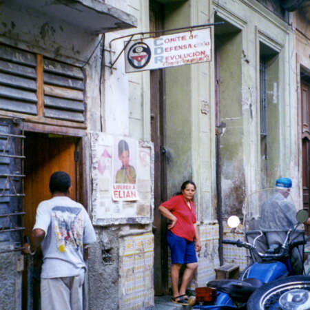 <p>A woman stands near an office for the Committee for the Defence of the Revolution. Set up in the early days of the Cuban revolution to counter saboteurs and counter-revolutionary activities, they have persisted as a sort of hybrid 'neighbourhood watch' and community welfare association. Critics contend they constitute little more than a vast network of informants for the government, while defenders claim they are essential for the local coordination of everything from vaccination campaigns and blood drives to hurricane evacuations.</p>
